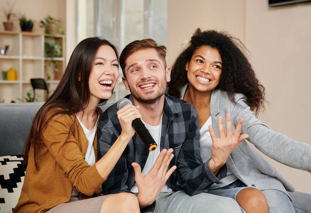 Τραγουδιστές καραόκε Τρεις χαμογελαστοί φίλοι που δείχνουν χαρούμενοι παίζοντας καραόκε στο σπίτι, τραγουδώντας με μικρόφωνο, καθισμένοι στον καναπέ στο μοντέρνο διαμέρισμα - Φωτογραφία, εικόνα