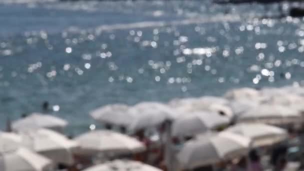 Fondo de verano desenfocado con sombrillas blancas y paisaje marino de playa borrosa con destellos de sol sobre agua ondulada - Imágenes, Vídeo