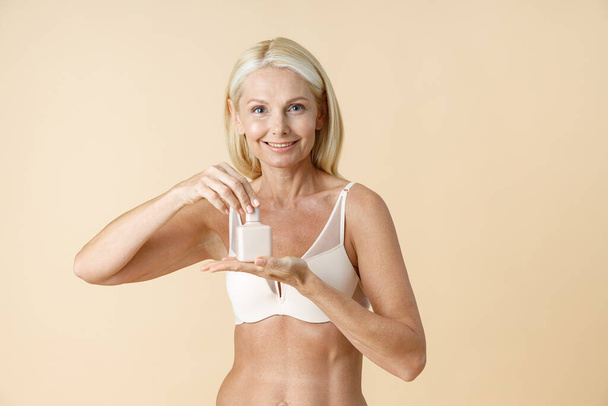 Adorable mature woman in underwear holding and opening white jar of skin care cream or lotion, standing isolated over beige background - Foto, imagen