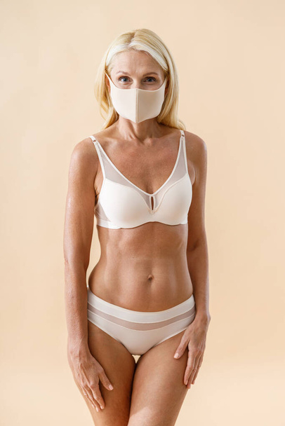 Studio shot of caucasian mature blonde woman wearing protective mask looking at camera while standing, posing in underwear isolated over beige background - Foto, imagen