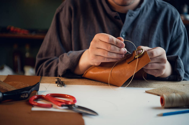 An einem Tisch in der Werkstatt sitzend näht ein junger Mann von Hand Lederelemente aneinander - Foto, Bild