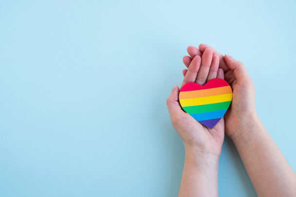 Naisten kädet pitävät sydäntä sateenkaaren väreissä vaaleansinisellä pohjalla. LGBT käsite, Pride kuukausi. Top näkymä, kopioi tilaa - Valokuva, kuva