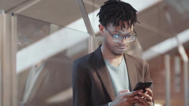 Waist up shot of young African American male office worker in eyeglasses and smart casual wear standing near glass wall and texting on smartphone during workday - Filmagem, Vídeo