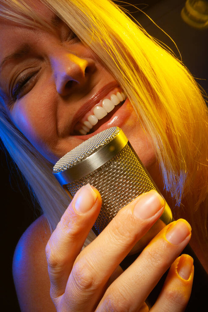 Vrouwelijke vocalist onder gelige verlichting zingt met passie in condensator microfoon. - Foto, afbeelding