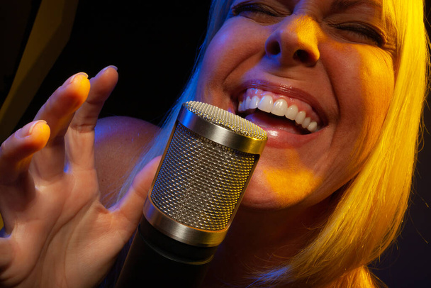 Γυναικείος τραγουδιστής κάτω από ζελέ φωτισμού τραγουδά με πάθος σε μικρόφωνο συμπυκνωτή. - Φωτογραφία, εικόνα