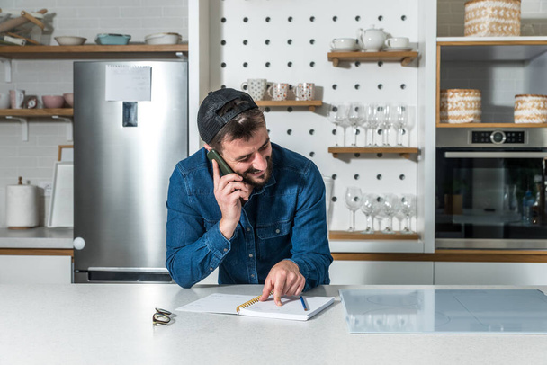 Young groom stands in the kitchen of his apartment and talking with his smartphone with friends he wants to invite to his bachelor party on the occasion of his wedding - Foto, Bild