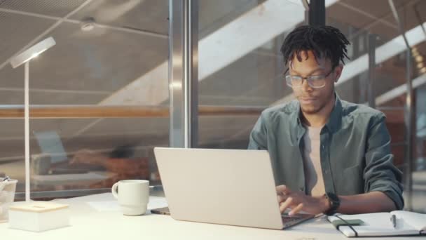 Plan de suivi du jeune homme afro-américain avec des textos de coiffure de tresse sur ordinateur portable et d'écrire les tâches dans le bloc-notes tout en travaillant au bureau moderne avec des murs de verre - Séquence, vidéo