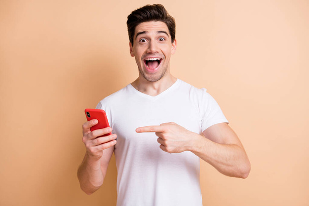 Photo of impressed funny guy open mouth direct finger phone unbelievable offer isolated on beige color background - Photo, image