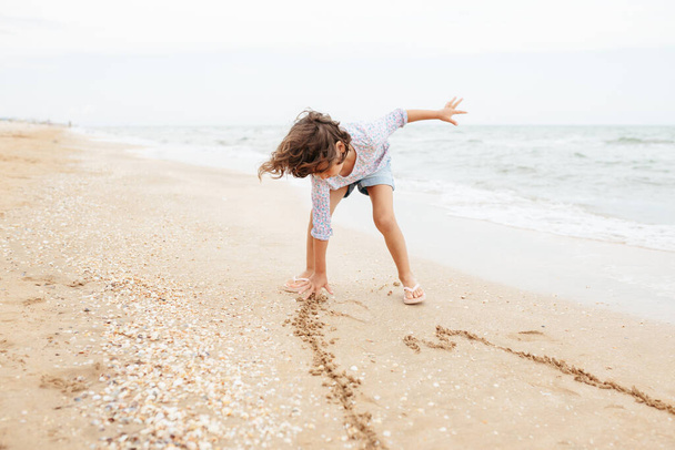 Five years cute curly caucasian girl painting on sand on the beach. - Fotó, kép