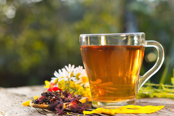 Bylinkový čaj v přírodě. Zátiší s bylinkovým čajem (černý čaj, ibiškový čaj, kalendula, hřebíček, kardomon, skořice) na rozmazaném pozadí. Připravený čaj, suchý čaj, okvětní lístky růží a heřmánek jsou na starém dřevěném stole. - Fotografie, Obrázek