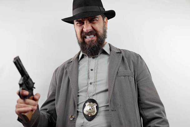 Detektyw z brodą w kapeluszu i odznaką policyjną trzymający broń ze wściekłym wyrazem twarzy. Koncepcja przestępstwa. - Zdjęcie, obraz