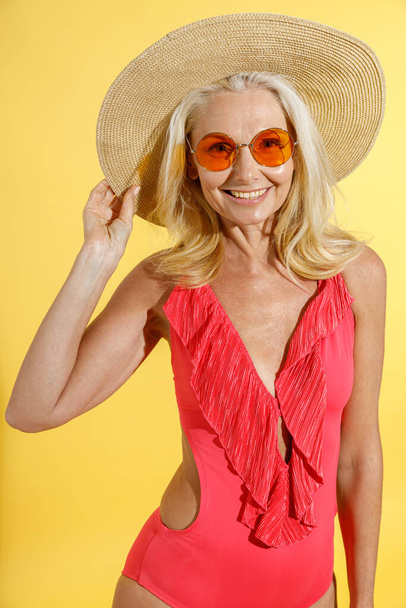 Studyjne ujęcie szczęśliwej blondynki w okularach przeciwsłonecznych, czerwonym stroju kąpielowym i słomkowym kapeluszu uśmiechniętej do kamery, ustawiającej kapelusz podczas pozowania, stojącej odizolowanej na żółtym tle - Zdjęcie, obraz