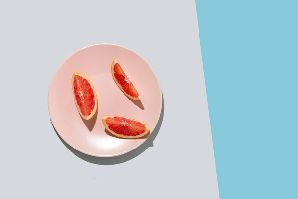 Tres trozos de rodajas de pomelo fresco en un plato de color rosa pastel sobre fondo gris y azul. Mínimo concepto de playa de verano con papeles. Piso con espacio de copia. - Foto, imagen