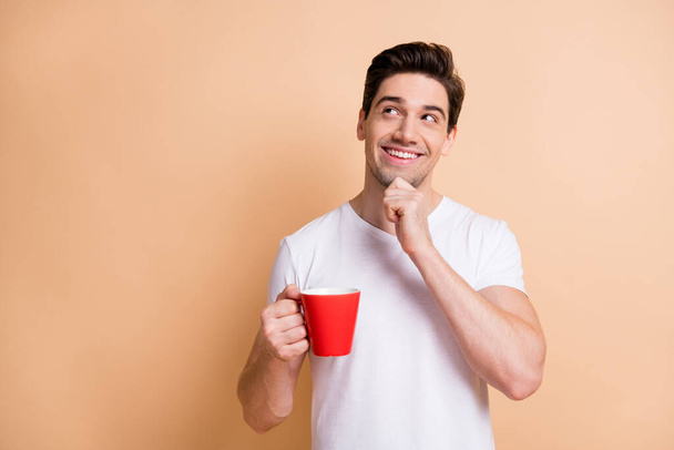 Portrait of cheerful handsome guy hand on chin look empty space hold coffee isolated on beige color background - Fotoğraf, Görsel