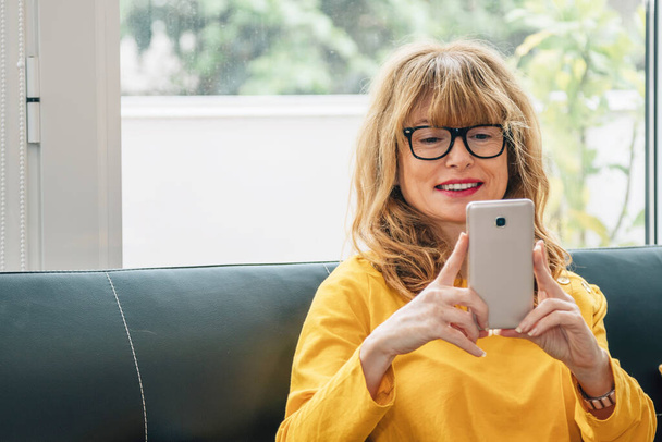 kobieta w domu uśmiechnięta patrząc na telefon komórkowy siedzący na kanapie - Zdjęcie, obraz