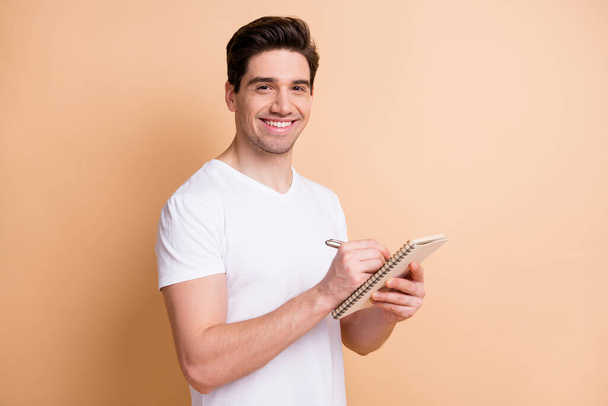 Portrait of half turned handsome guy toothy smile writing in textbook look camera isolated on beige color background - 写真・画像
