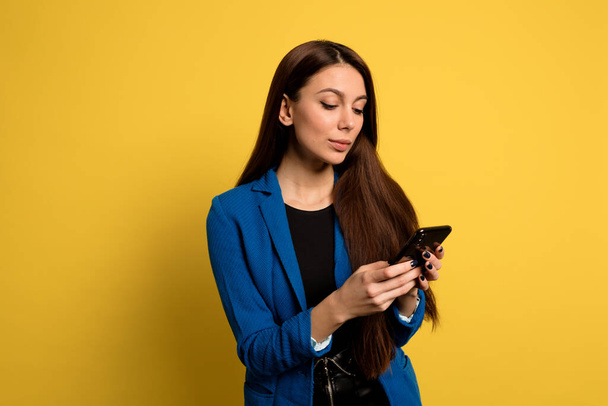 Γοητευτική όμορφη γυναίκα με μακριά σκούρα μαλλιά κύλιση smartphone πάνω από κίτρινο φόντο - Φωτογραφία, εικόνα