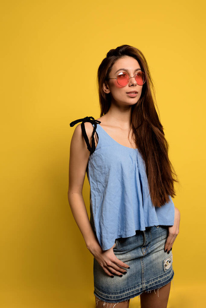 Stylish pretty woman wearing blue t-shirt and denim skirt and round pink glasses posing over yellow backdrop - Foto, afbeelding