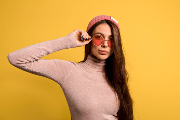 Stylish modern woman with long straight hair wearing pink shirt and cap touching her round pink glasses and smiling  - Fotografie, Obrázek