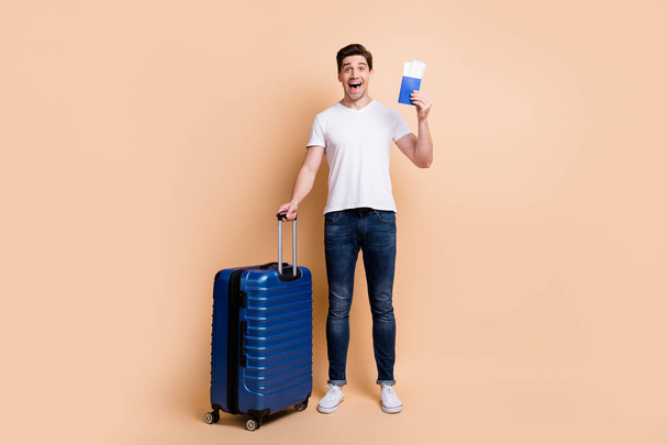 Full size photo of impressed brunet man stand hold tickets bag wear t-shirt jeans sneakers isolated on beige background - 写真・画像