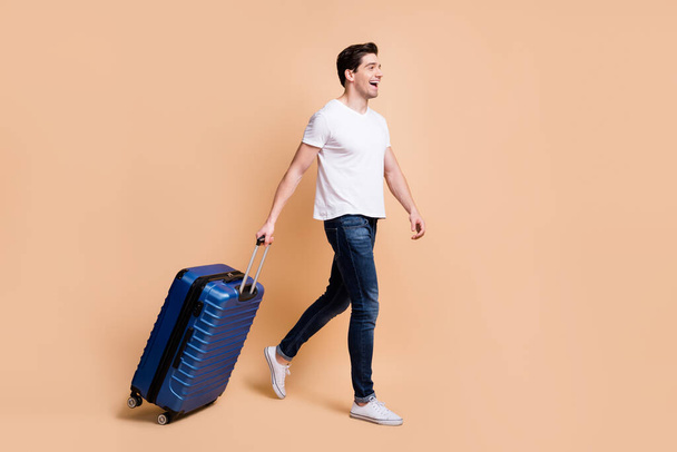 Full size profile photo of impressed brunet man go hold bag wear t-shirt jeans sneakers isolated on beige background - 写真・画像