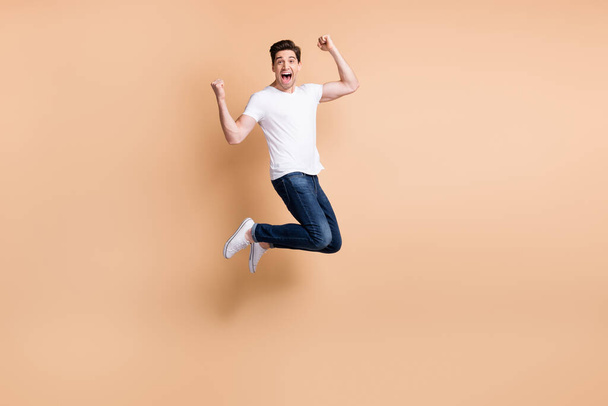 Full size profile photo of hooray brunet man jump yell wear t-shirt jeans sneakers isolated on beige background - 写真・画像