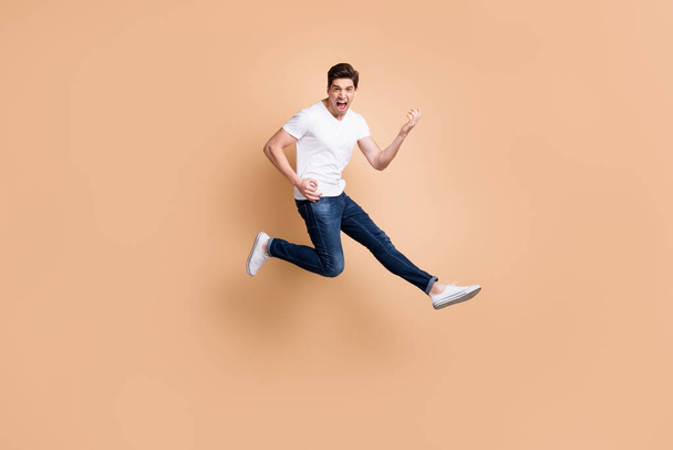 Full size profile photo of hooray brunet man jump play guitar yell wear t-shirt jeans sneakers isolated on beige color background - Foto, Imagen