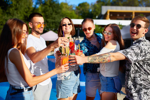 Group of friends having fun at poolside summer party clinking glasses with summer cocktails on sunny day near swimming pool. People toast drinking fresh juice at luxury villa on tropical vacation. - 写真・画像