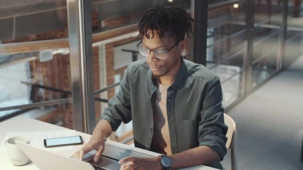 Hochwinkelaufnahme eines afroamerikanischen männlichen Büroangestellten, der am Laptop tippt, während er am Schreibtisch im zweiten Stock des modernen Business Centers mit Loft-Interieur arbeitet - Filmmaterial, Video