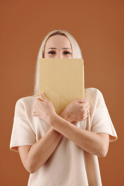 Portret wesołej młodej kobiety przykrywającej twarz tabletem i patrzącej w kamerę - Zdjęcie, obraz