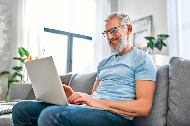 A mature man is sitting on the couch with a laptop on his lap at home. Freelance, work online. The man is texting on his laptop. - Foto, imagen