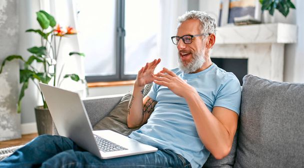 Een volwassen man zit thuis op de bank met een laptop op schoot en praat op een videogesprek. Blijf thuis concept. Freelance, thuiswerk. - Foto, afbeelding