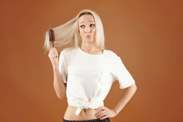 Muito loira jovem escovando seu cabelo seco emaranhado, isolado em marrom - Foto, Imagem