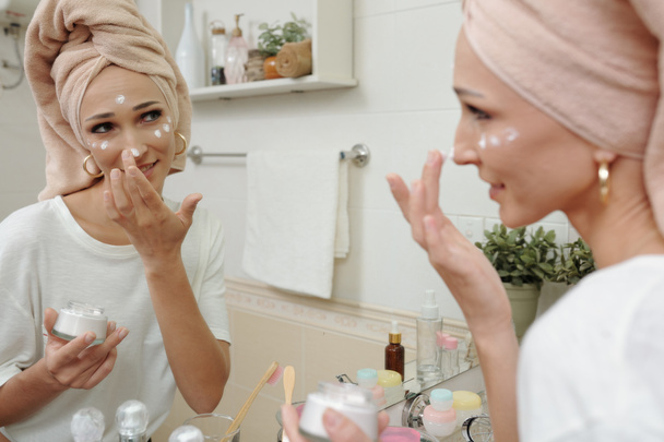 Jolie jeune femme souriante debout au miroir de salle de bains et appliquant la crème raffermissante de collagène de réparation avec des peptides après la douche du matin - Photo, image