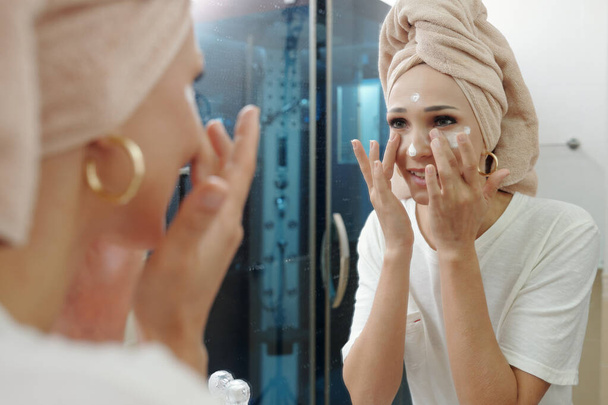 Улыбающаяся молодая женщина смотрит в зеркало, нанося увлажняющий крем против старения на лицо - Фото, изображение