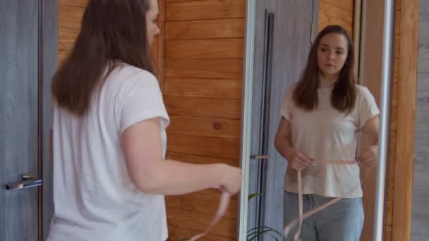 Portret van een vrouw van middelbare leeftijd die haar taille meet met tape voor de spiegel thuis - Video