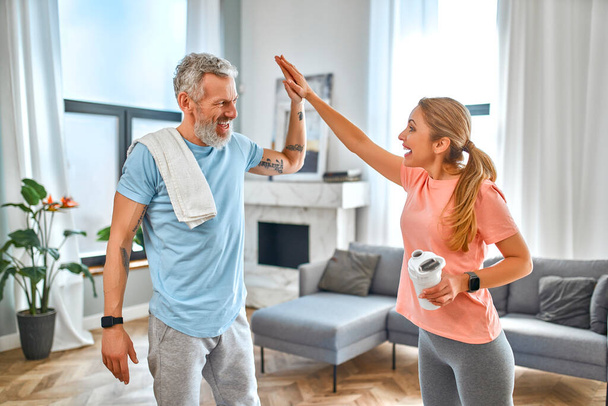 Sport, fitness en een gezonde levensstijl concept. Volwassen echtpaar geven vijf gebaar en doen oefeningen thuis en veel plezier. Hoe gezond te blijven in quarantaine. - Foto, afbeelding