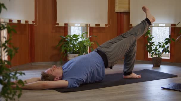 Fit hombre caucásico haciendo ejercicio de yoga estirando los músculos de las piernas y los abdominales usando la computadora en casa - Imágenes, Vídeo