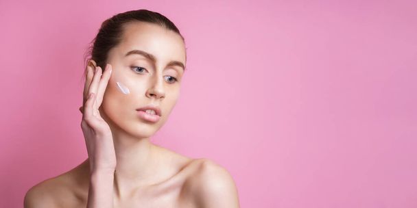 Beautiful young woman applying cream on her face against color background - Foto, imagen