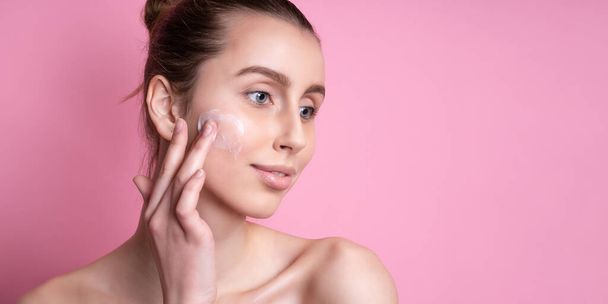 Beautiful young woman applying cream on her face against color background - Фото, изображение