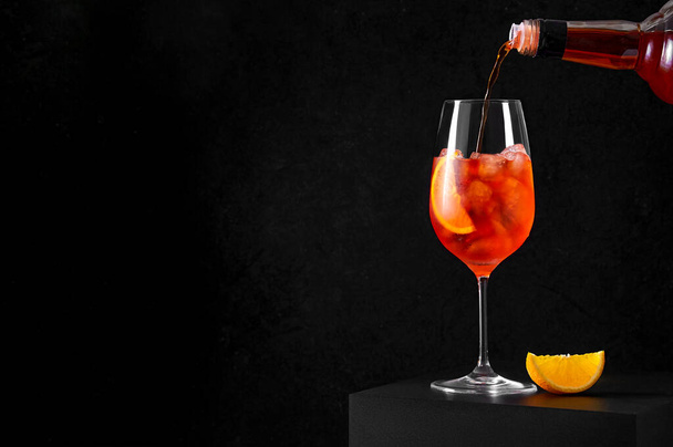 Verser Aperol Spritz Cocktail dans un verre à vin avec de la glace et une tranche d'orange sur fond sombre. Longue boisson gazeuse - Photo, image