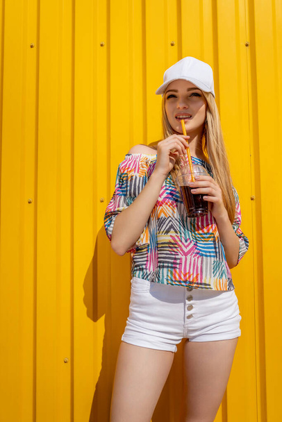 Pretty young hipster style girl have fun outdoor in white cap and eating ice cream on yellow background urban style - Фото, изображение