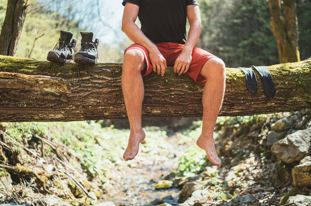  Man sitting on the fallen tree log over the mountain forest stream while he waiting for socks laundry drying and trekking boots. Active people traveling, hiking or trekking concept image. - Zdjęcie, obraz