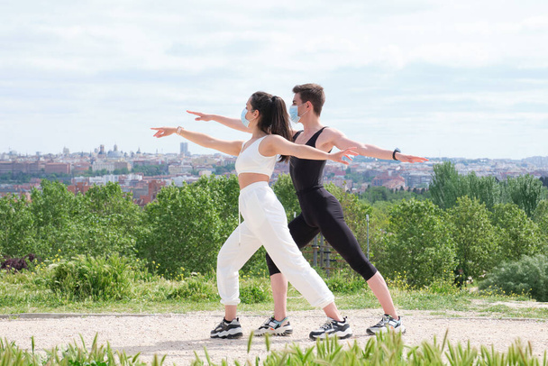 Young couple doing warrior two yoga pose in a park wearing protective face masks. Fitness outside during pandemic. - Foto, Imagen