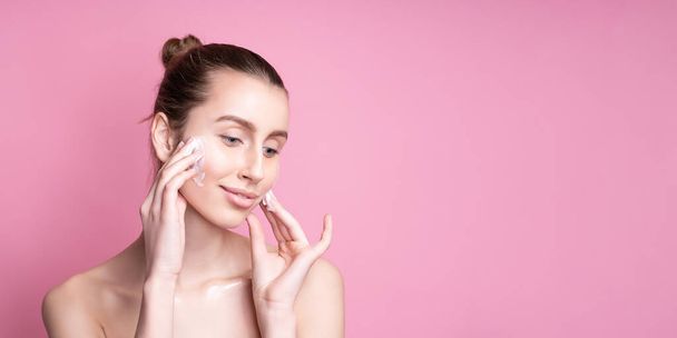 Beautiful young woman applying cream on her face against color background - Photo, image