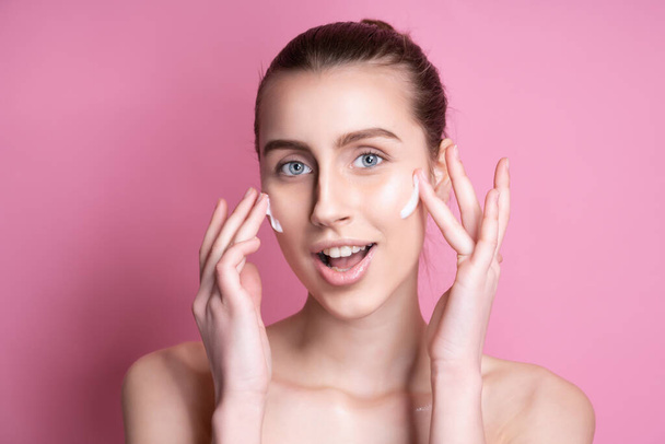 Beautiful young woman applying cream on her face against color background - Foto, afbeelding
