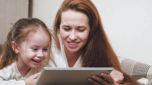 Μια ευτυχισμένη οικογένεια, η μαμά και ένα παιδί ψάχνουν σε μια ενδιαφέρουσα γελοιογραφία στην οθόνη tablet και γέλιο, η ζωή είναι σε απευθείας σύνδεση μάθηση στην εφαρμογή, μαμά και η κόρη βρίσκονται στον καναπέ και - Φωτογραφία, εικόνα