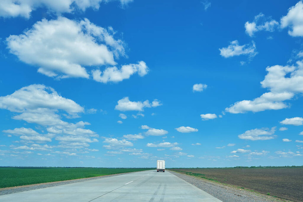 Αγροτικό τοπίο με τσιμεντένιο δρόμο και φορτηγό μακριά σε κίνηση. Πράσινα πεδία στο παρασκήνιο. Ηλιόλουστη ανοιξιάτικη μέρα με μπλε ουρανό και άσπρα σύννεφα. - Φωτογραφία, εικόνα