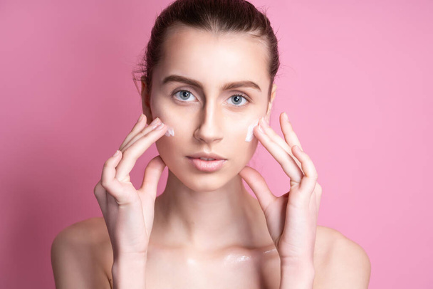 Beautiful young woman applying cream on her face against color background - Photo, Image