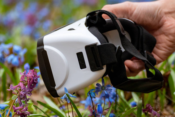 Mano sosteniendo gafas de realidad virtual 3D en primavera flores silvestres azules. VR tecnología de engranajes gafas vista lateral de cerca. Sistema de entretenimiento óptico para smartphones - Foto, Imagen
